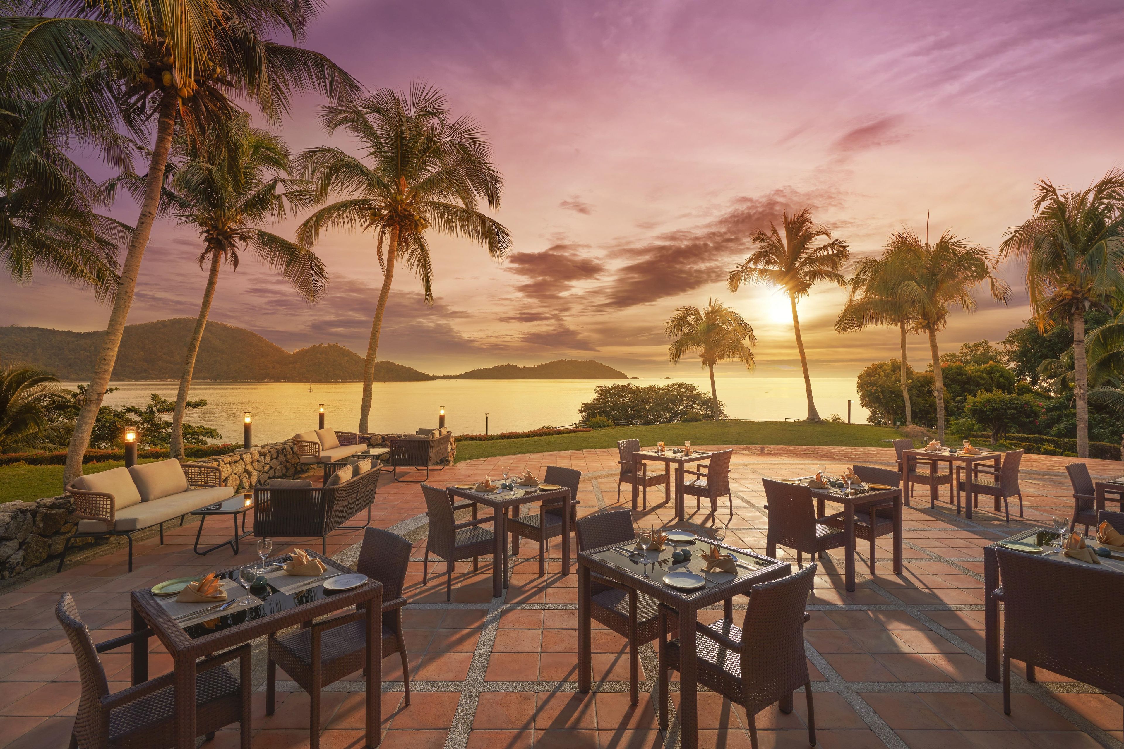 An outdoor dining area with tables and chairs, palm trees, and a view of the water and mountains.
