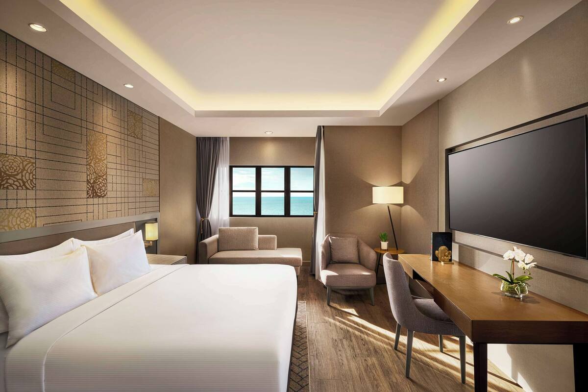Ein Hotelzimmer mit einem großen Bett, einem Schreibtisch mit Stuhl, einem Fernseher und Meerblick durch das Fenster.