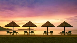 Een strand met vier rieten parasols en ligbedden gericht op de zee bij zonsondergang.