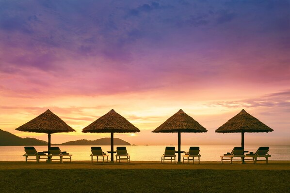 Una playa con cuatro sombrillas de paja y sillones de descanso frente al mar al atardecer.