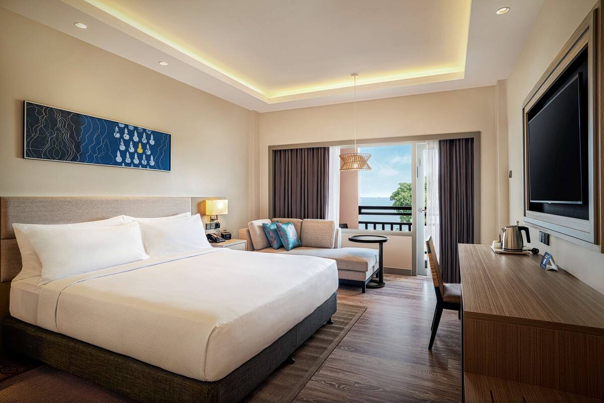 Ein Hotelzimmer mit einem großen Bett, einem Schreibtisch, einem Stuhl, einem kleinen Tisch, einem Fernseher und einem Balkon mit Blick.