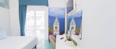 A hotel room with a bed, a desk with a plant, a chair, and a large window with a view of a coastal landscape.