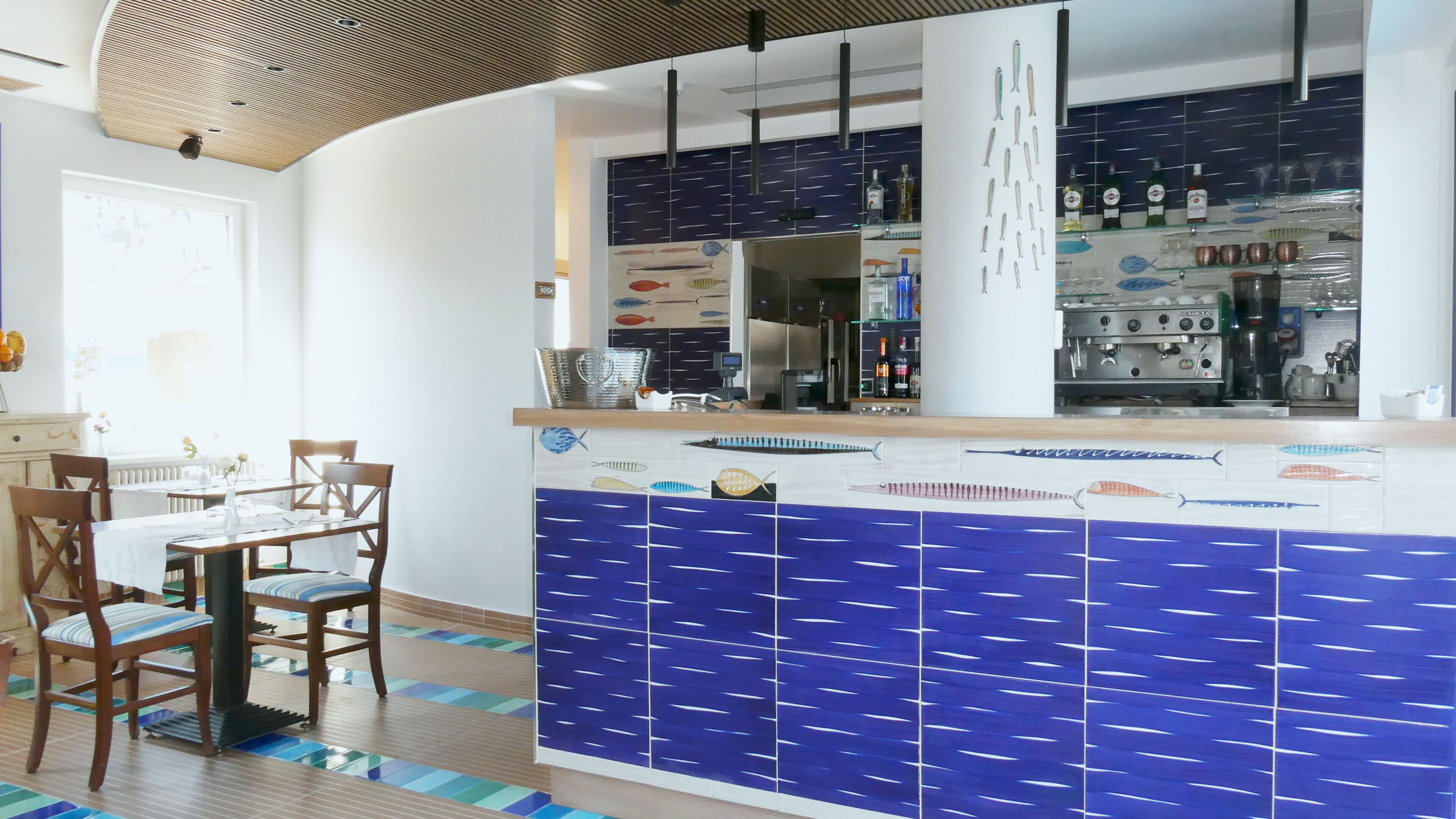 Un restaurante con una pared de azulejos con motivos de peces azules, sillas de madera, una mesa blanca y una barra con varias botellas.