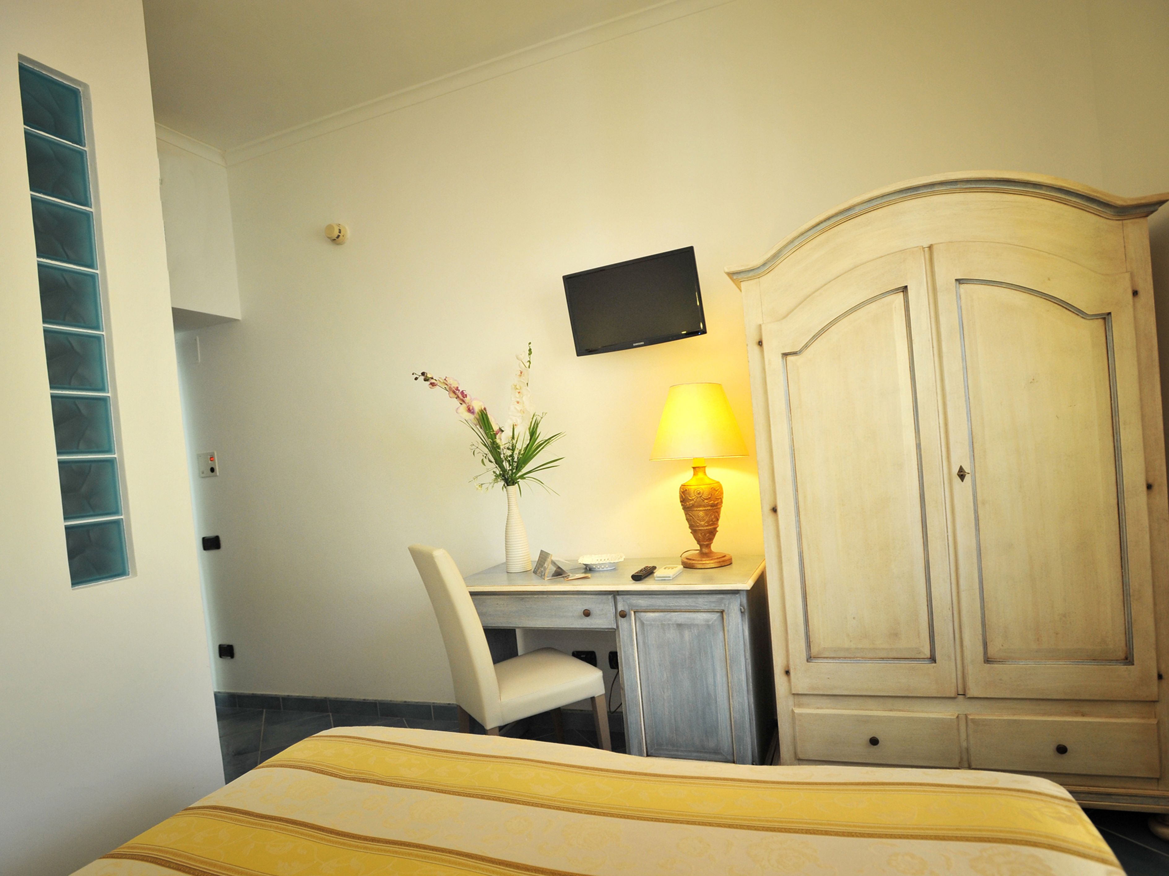 A hotel room with a bed, a desk with a chair, a lamp, a TV mounted on the wall, and a wardrobe.