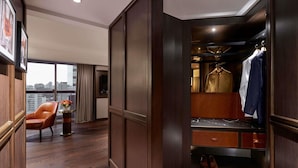 Habitación de hotel con piso de madera, zona de estar con una silla y una mesita, armario con ropa colgada y vista de la ciudad a través de amplios ventanales.