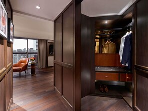 Hotelkamer met houten vloer, zithoek met stoel en klein tafeltje, kledingkast met daarin hangende kleding, en stadszicht door grote ramen.