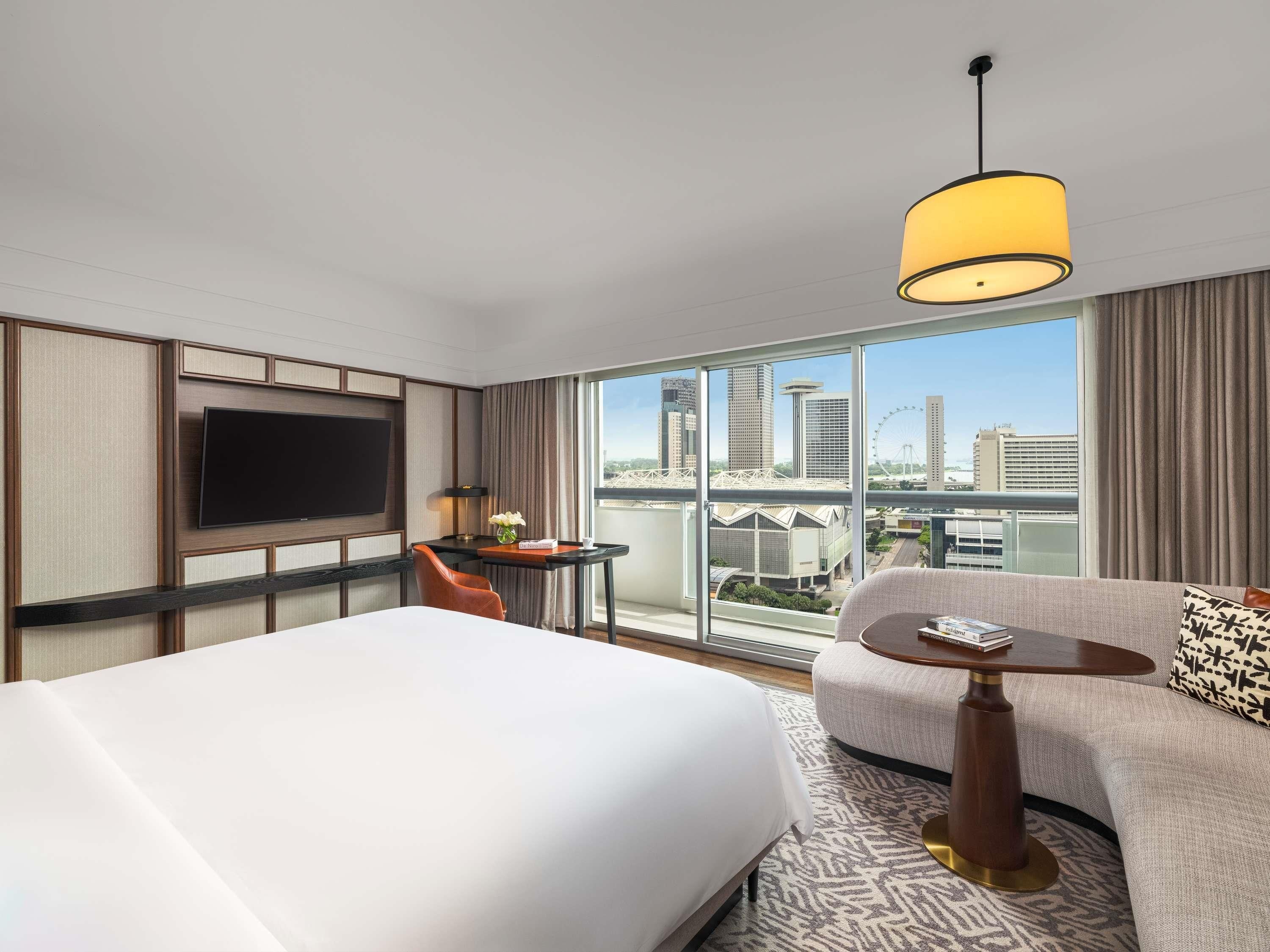 A hotel room with a large bed, a sofa, a small table, a TV, and a city view through large windows.