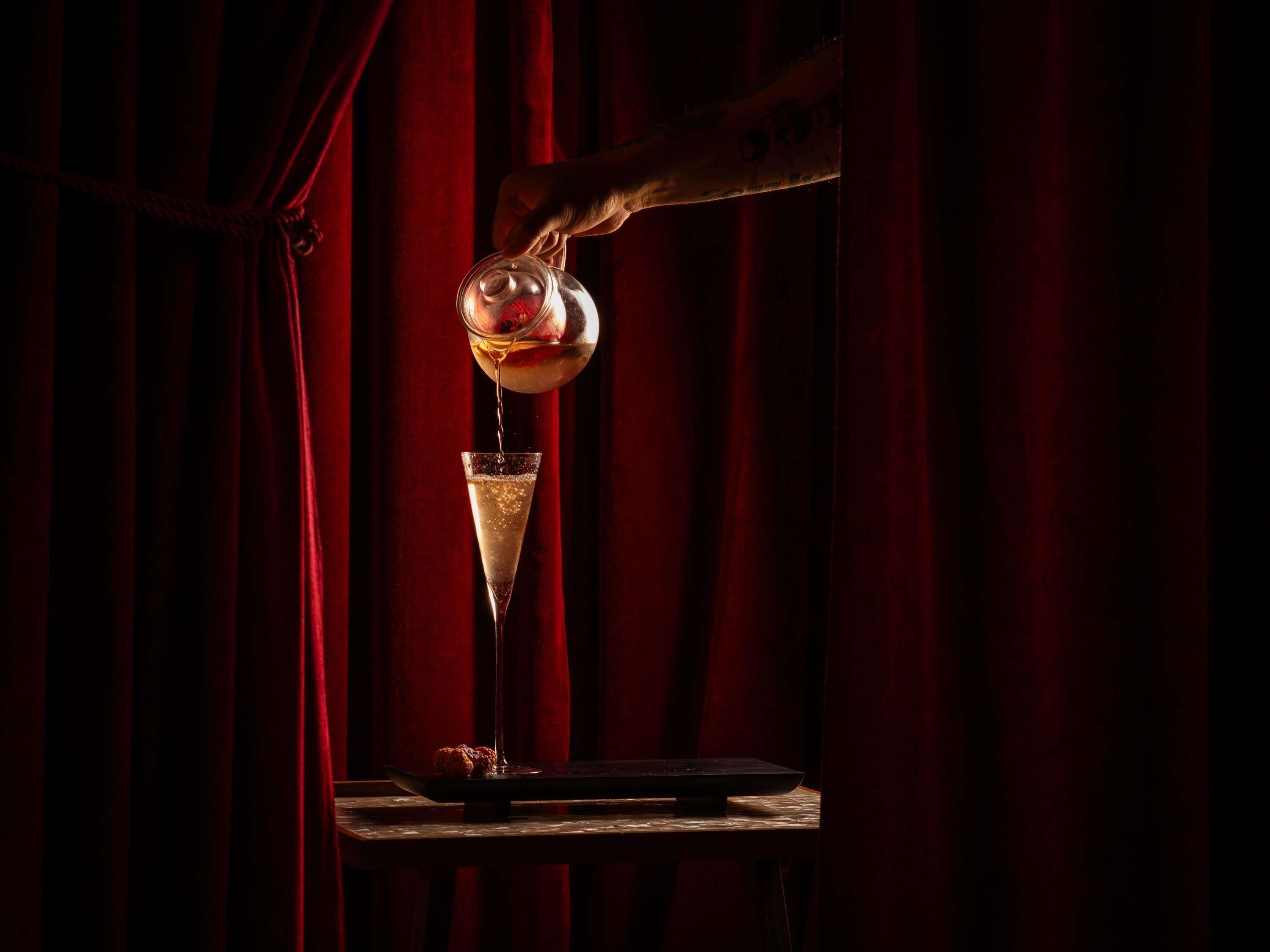 A hand pouring a drink into a glass on a table with a red curtain backdrop.