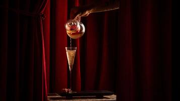 A hand pouring a drink into a glass on a table with a red curtain backdrop.