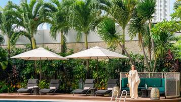 Zona de piscina con sillas de descanso, sombrillas y una persona con un vestido blanco.