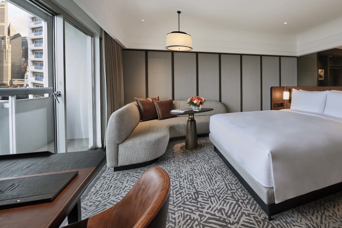 Een moderne hotelkamer met een groot bed, een bank, een tafeltje met een bloevas, en uitzicht op de stad door het raam.