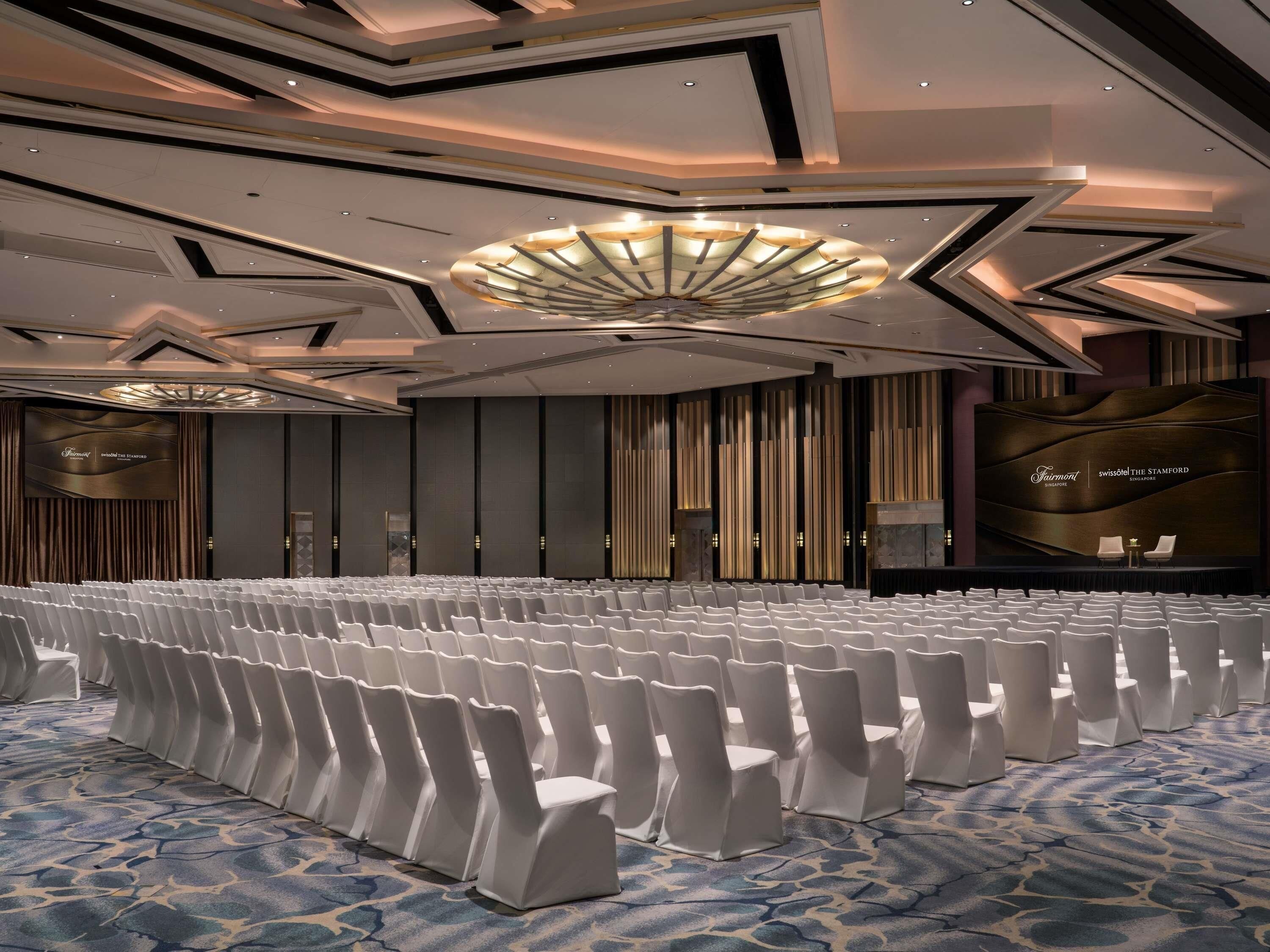 Un salón de conferencias con filas de sillas blancas, una pantalla grande y un diseño moderno en el techo.