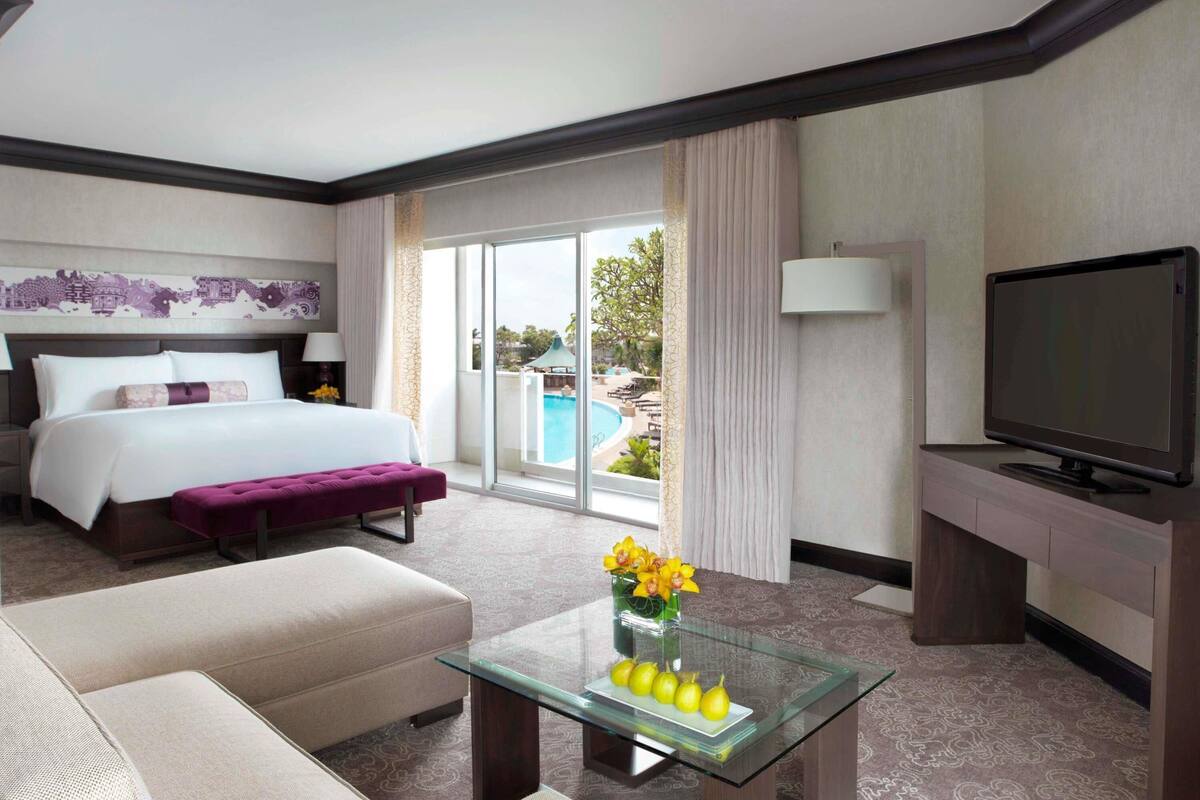 A hotel room with a bed, a sofa, a glass coffee table with fruit and flowers, a TV, and a view of a pool and greenery.