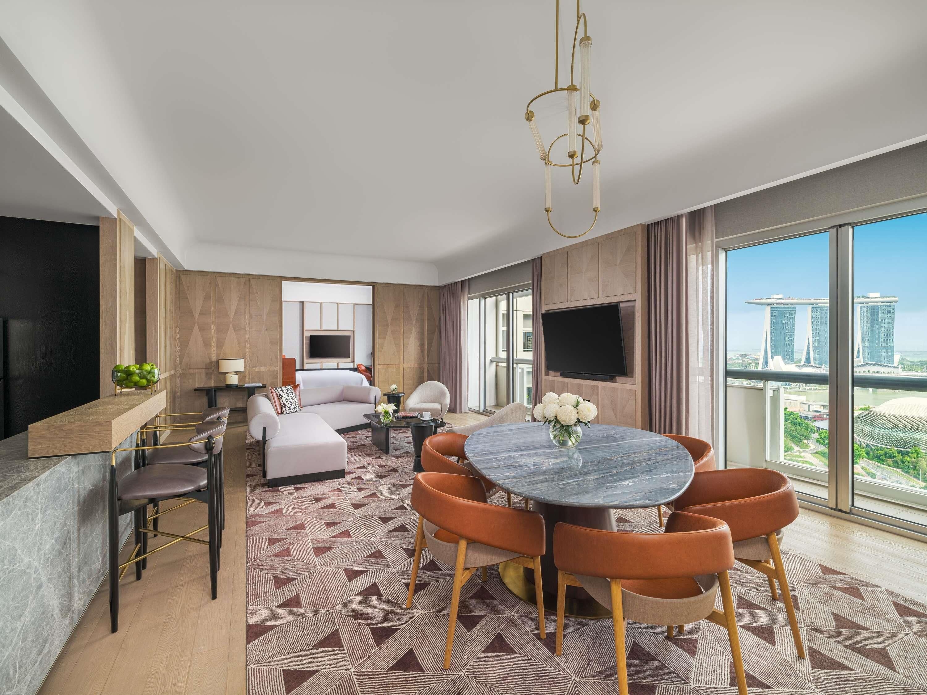 A modern hotel room with a large bed, a TV, a dining area with a round table, and a view of a city skyline.