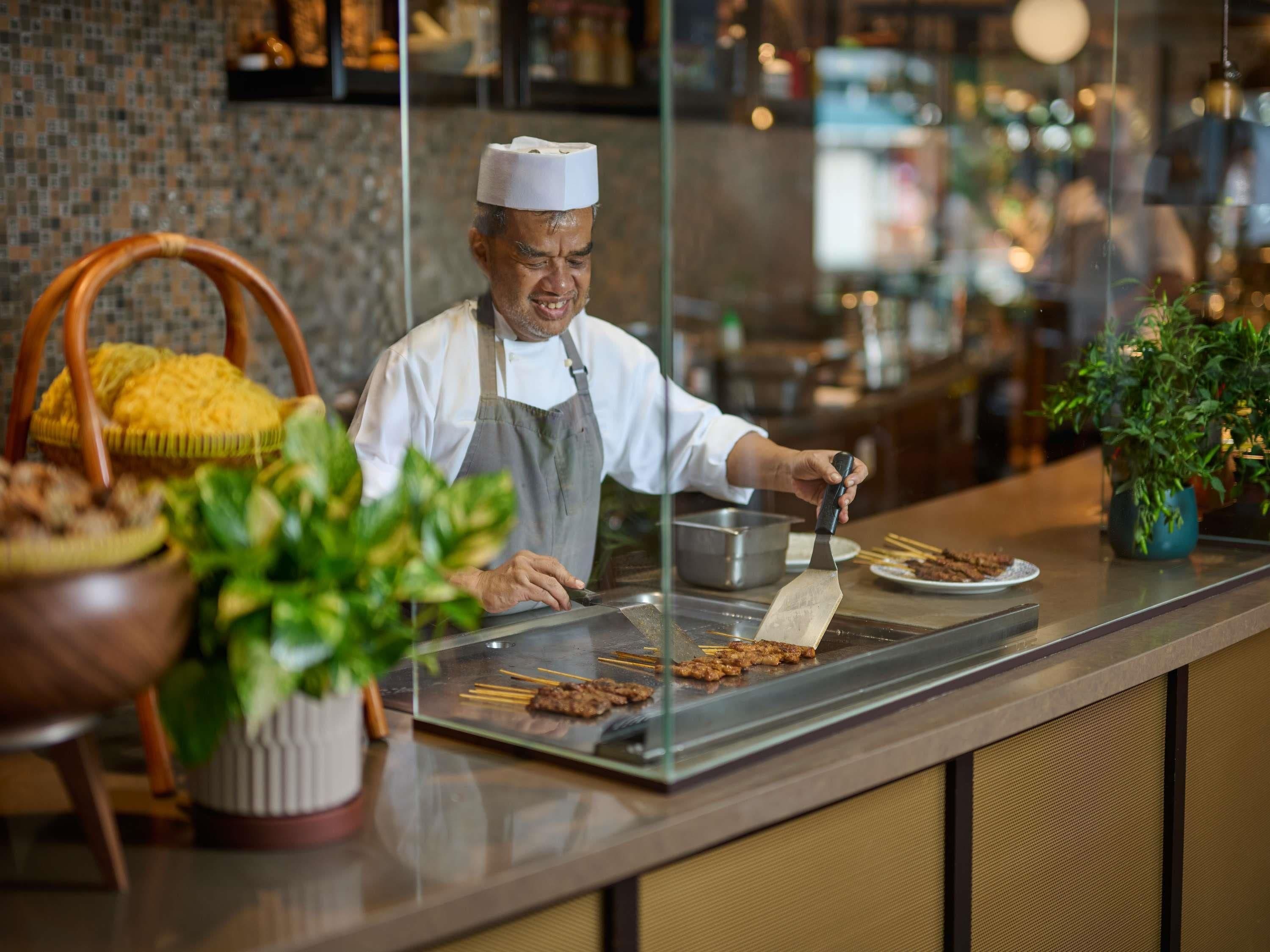 A chef preparing food at a counter with a grill, a basket of yellow corn, and potted plants.