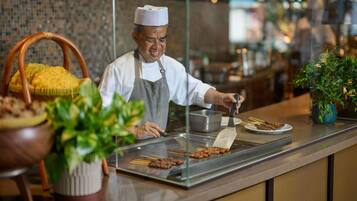 A chef preparing food at a counter with a grill, a basket of yellow corn, and potted plants.
