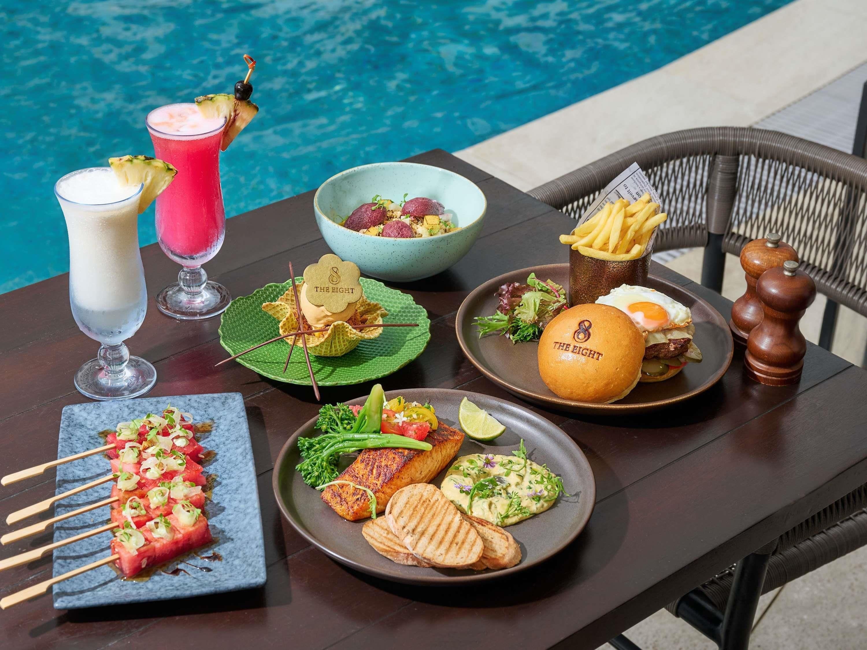 Mesa de jantar com diversos pratos, incluindo espetinhos, salmão grelhado e um hambúrguer, com vista para a piscina.
