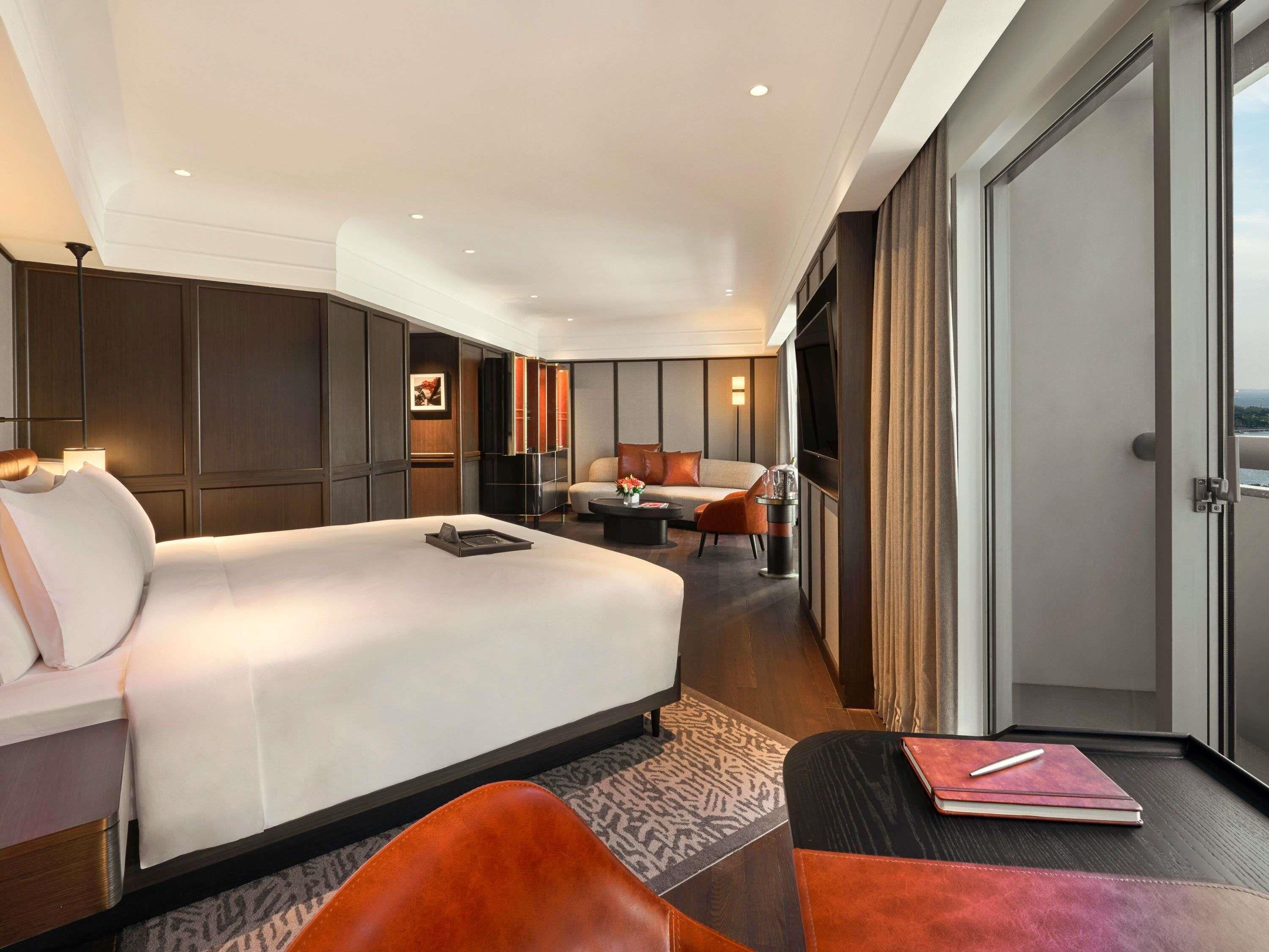 A modern hotel room with a large bed, a seating area, a TV, and a view of the outdoors.