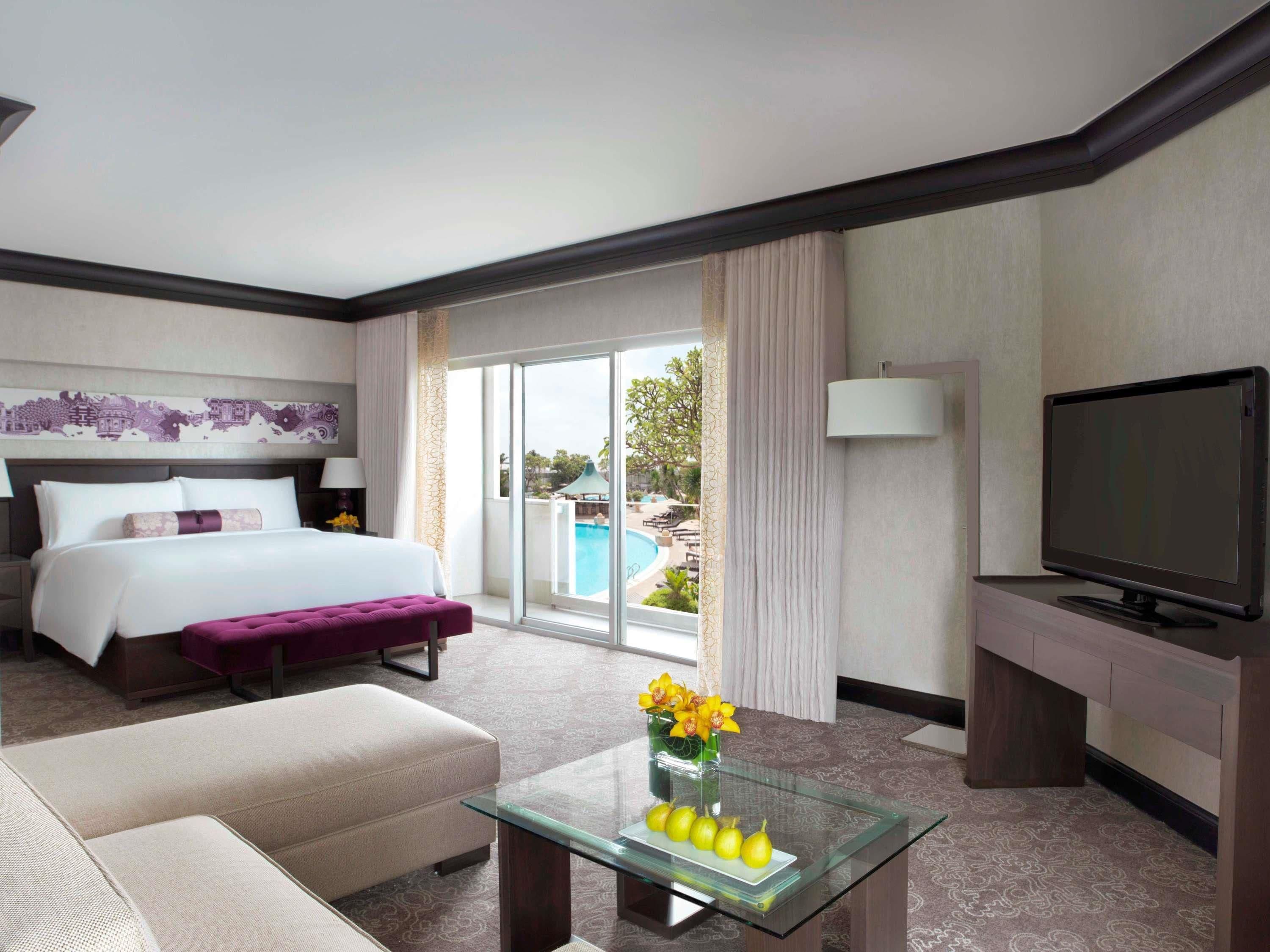 A hotel room with a bed, a sofa, a glass coffee table with fruit and flowers, a TV, and a view of a pool and greenery.
