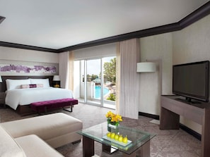 A hotel room with a bed, a sofa, a glass coffee table with fruit and flowers, a TV, and a view of a pool and greenery.