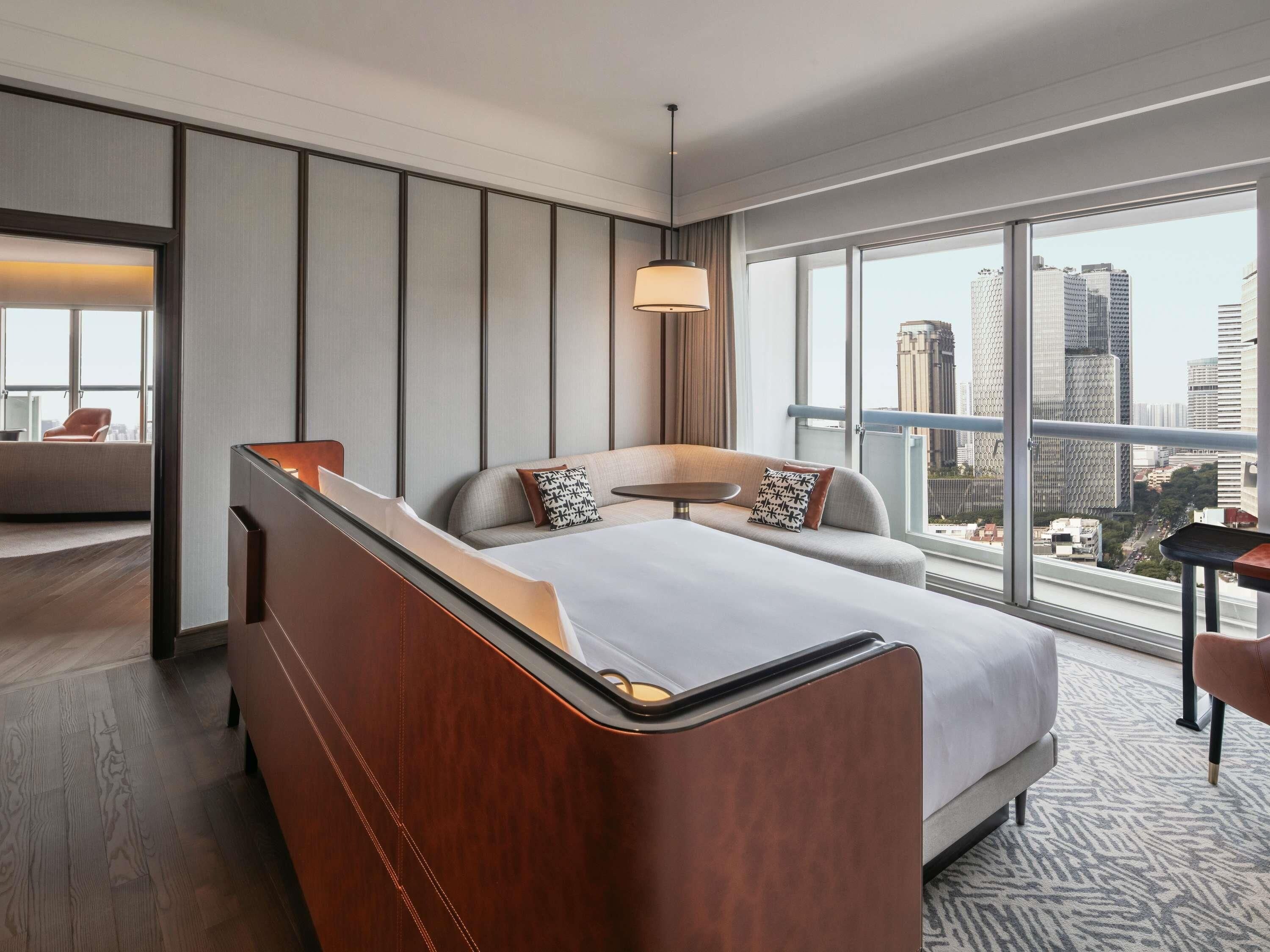 A modern hotel room with a large bed, a sofa, a small table, and a view of the cityscape through large windows.