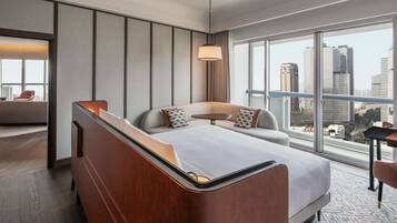 A modern hotel room with a large bed, a sofa, a small table, and a view of the cityscape through large windows.