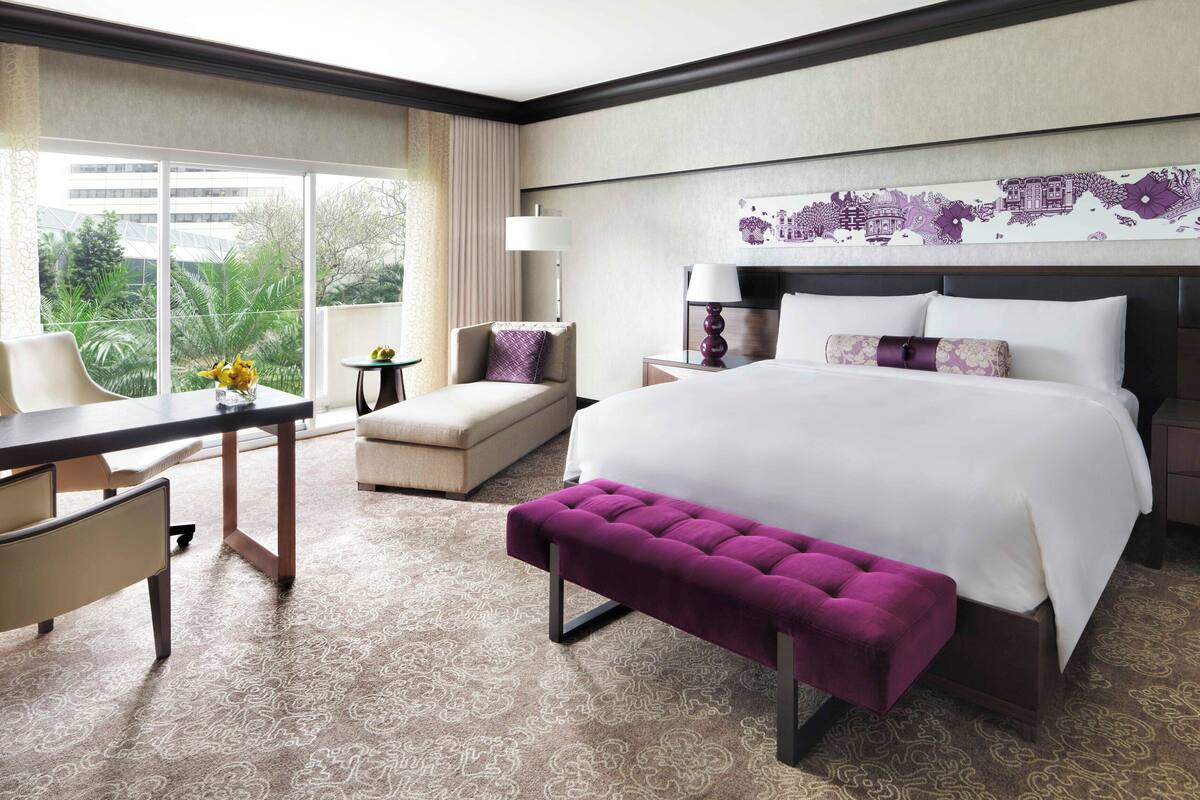 A hotel room with a large bed, a desk, a chair, a small table, and a view of greenery through the window.