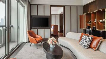 A modern living room with a sofa, armchair, and coffee table, a TV mounted on the wall, and a view through large windows.
