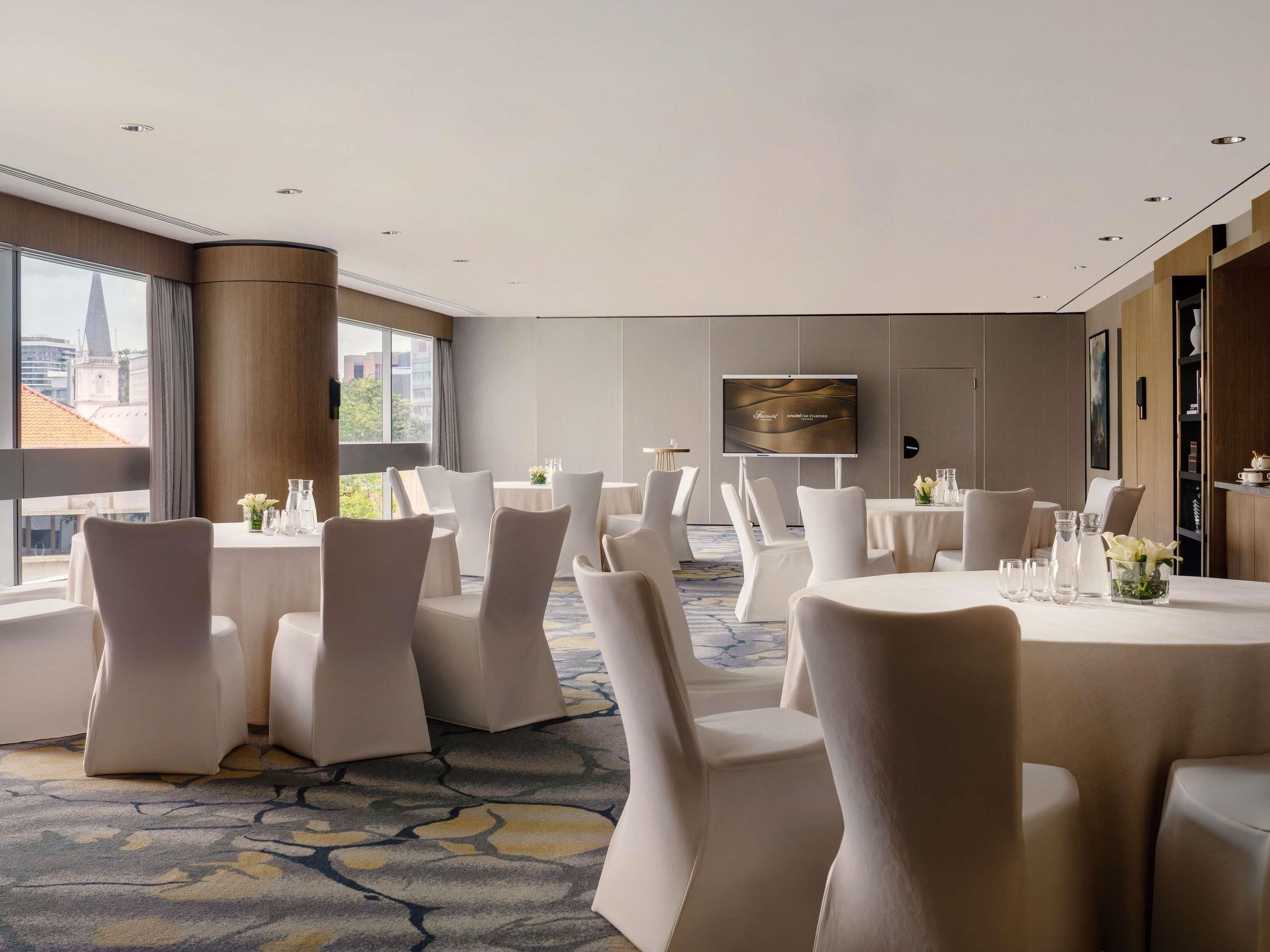 A hotel conference room with round tables, white chairs, a large screen, and a city view.