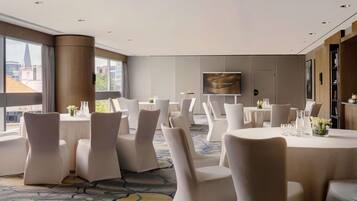 A hotel conference room with round tables, white chairs, a large screen, and a city view.