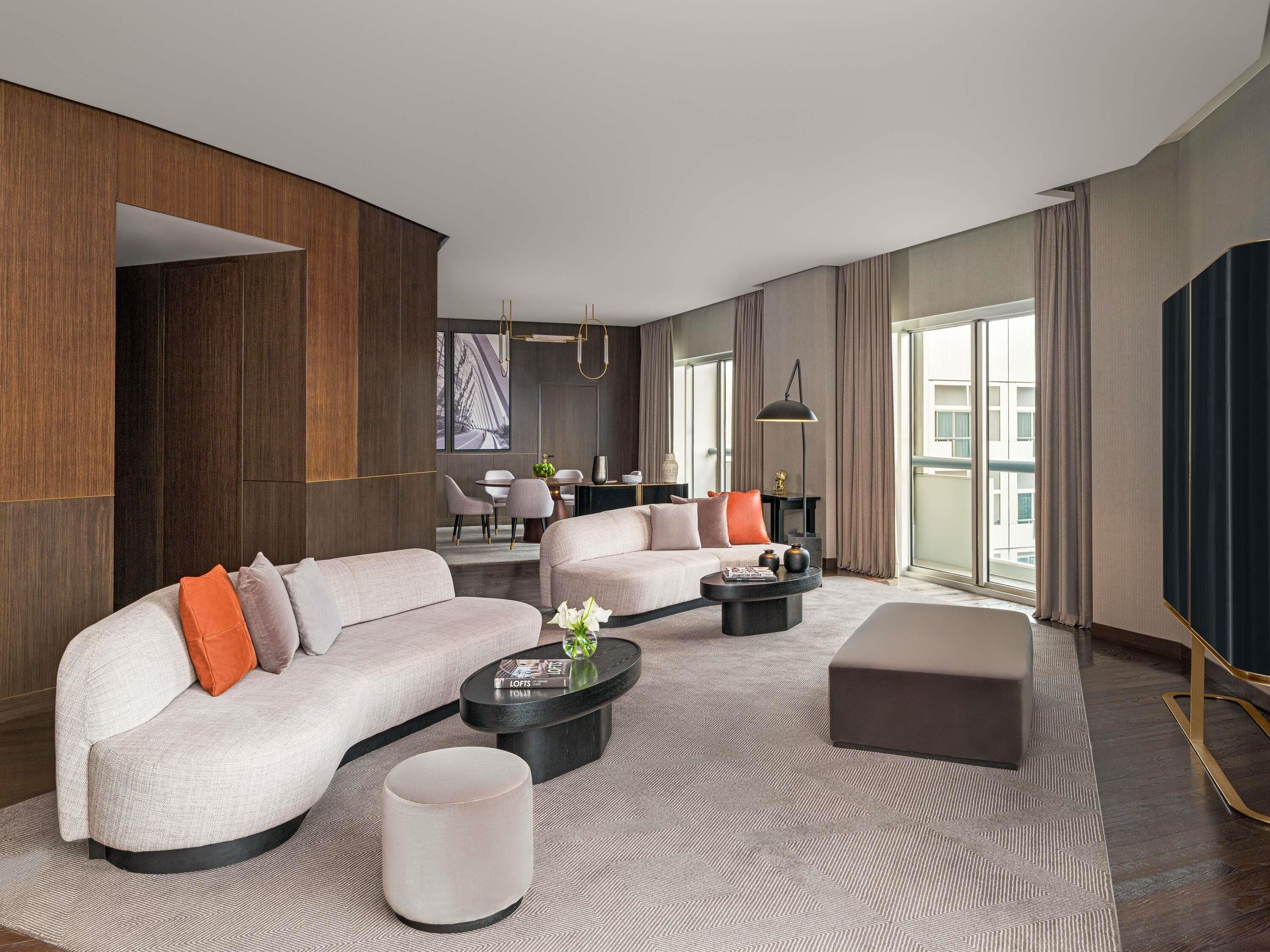 A modern hotel room with a sectional sofa, a round coffee table, a small side table, and a large window offering a view of the city.