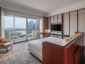 Habitación de hotel con una cama grande, televisión, escritorio y una silla. Se ve un paisaje urbano a través de amplios ventanales.