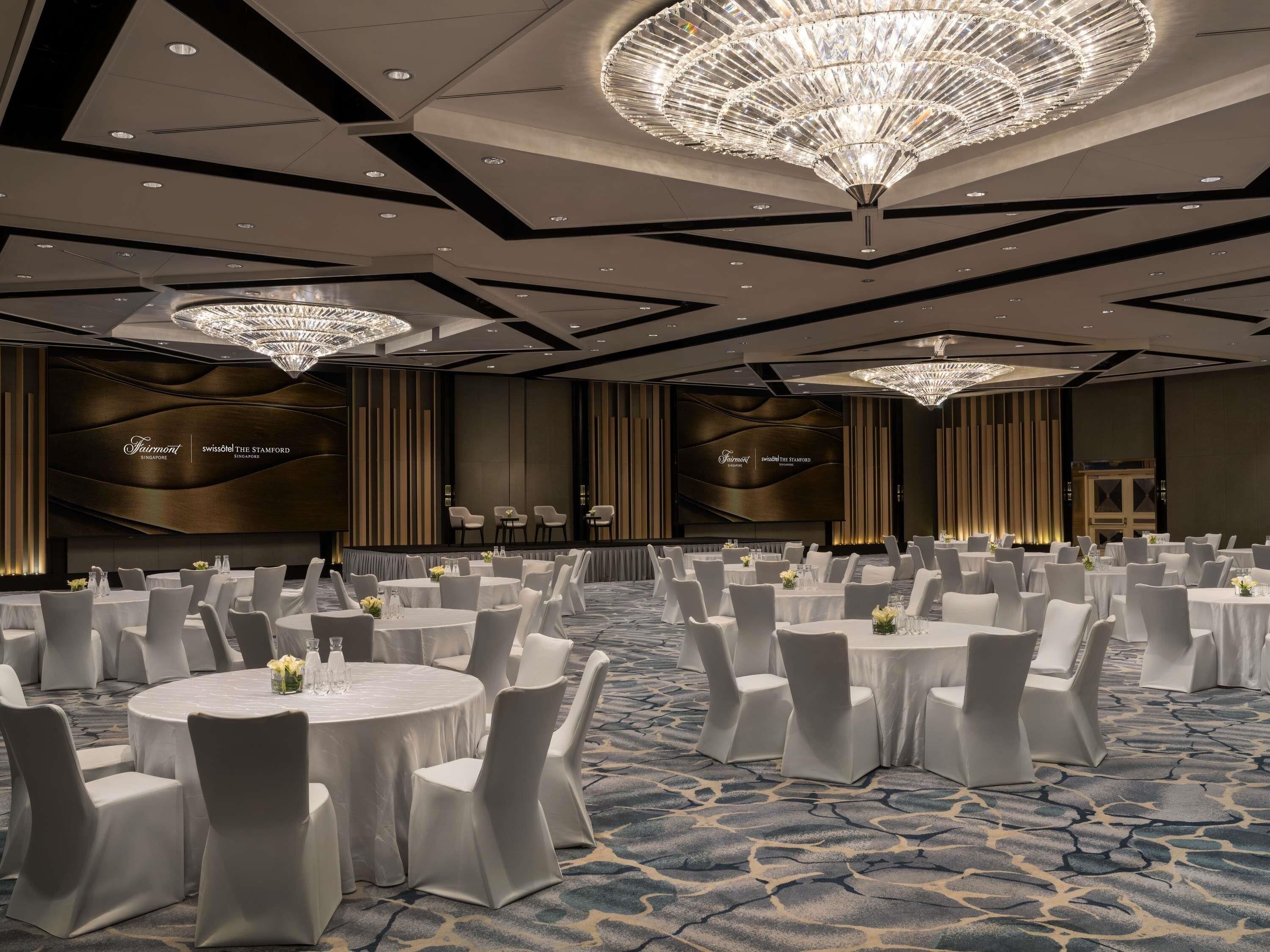 A banquet hall with round tables, white chairs, and chandeliers.