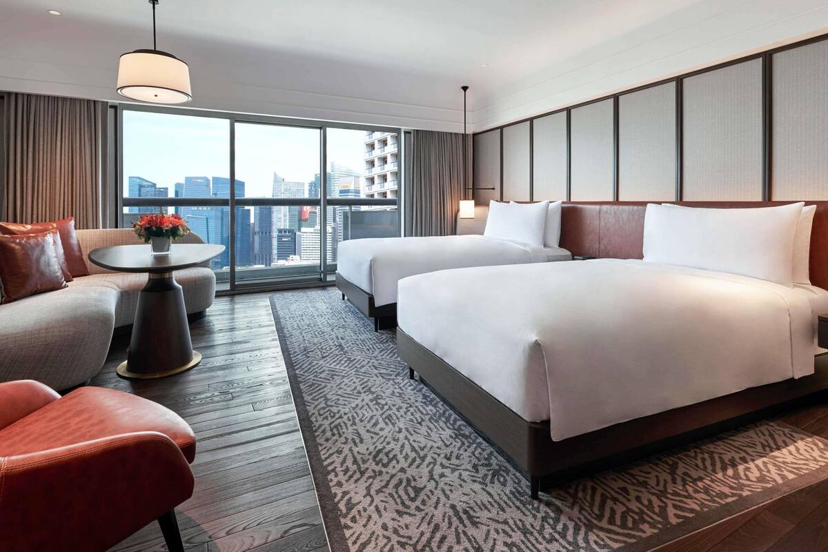 A hotel room with two beds, a sofa, a small table with a vase, and a large window offering a city view.