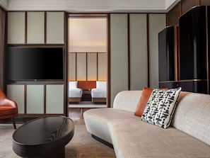 A modern hotel room with a large flat-screen TV, a sofa with patterned cushions, a leather armchair, and a round coffee table.