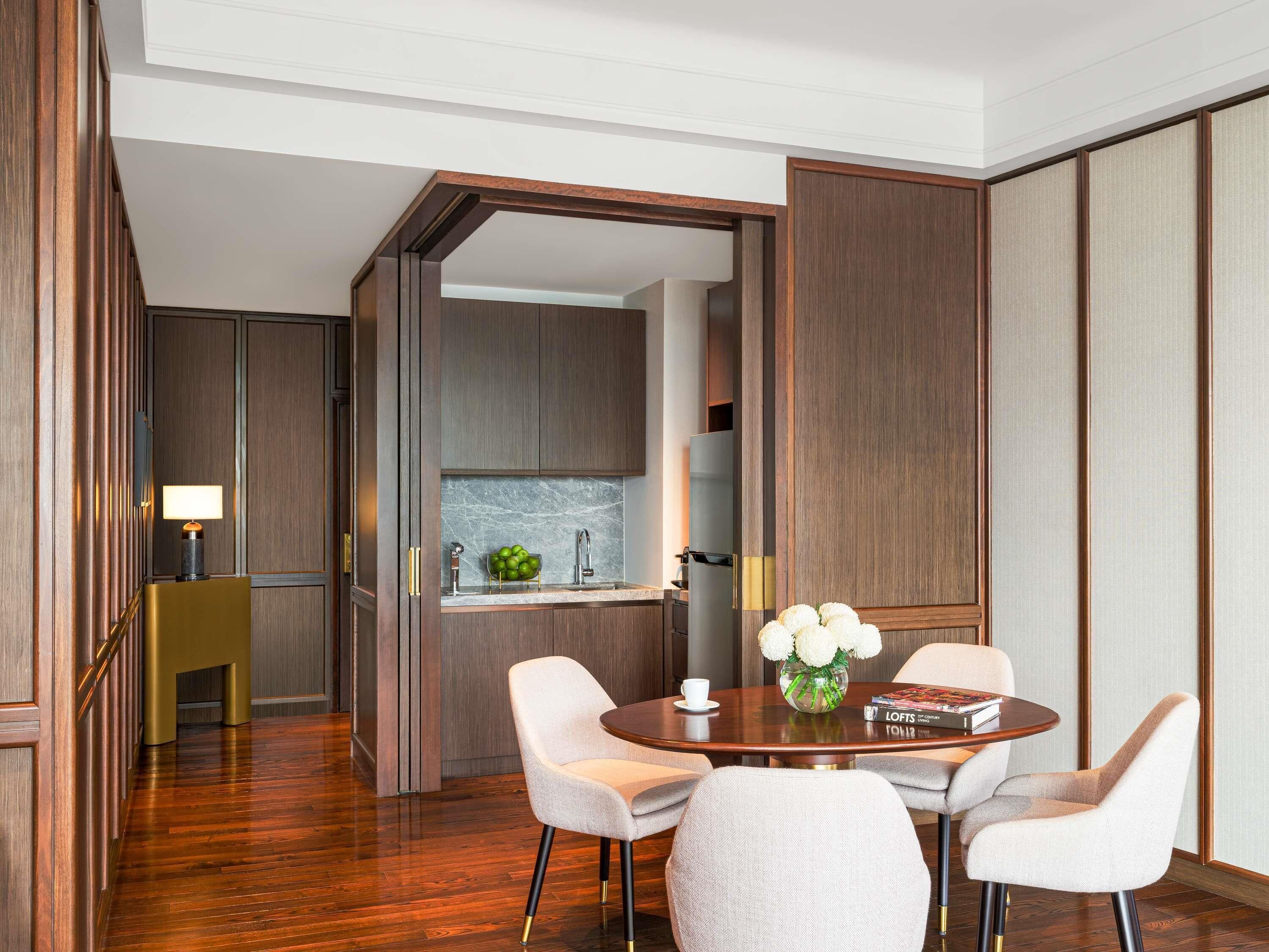 Een moderne hotelkamer met een houten vloer, een zithoek met een ronde tafel en vier stoelen, een keuken met een spoelbak en kastjes, en uitzicht door een open deur.