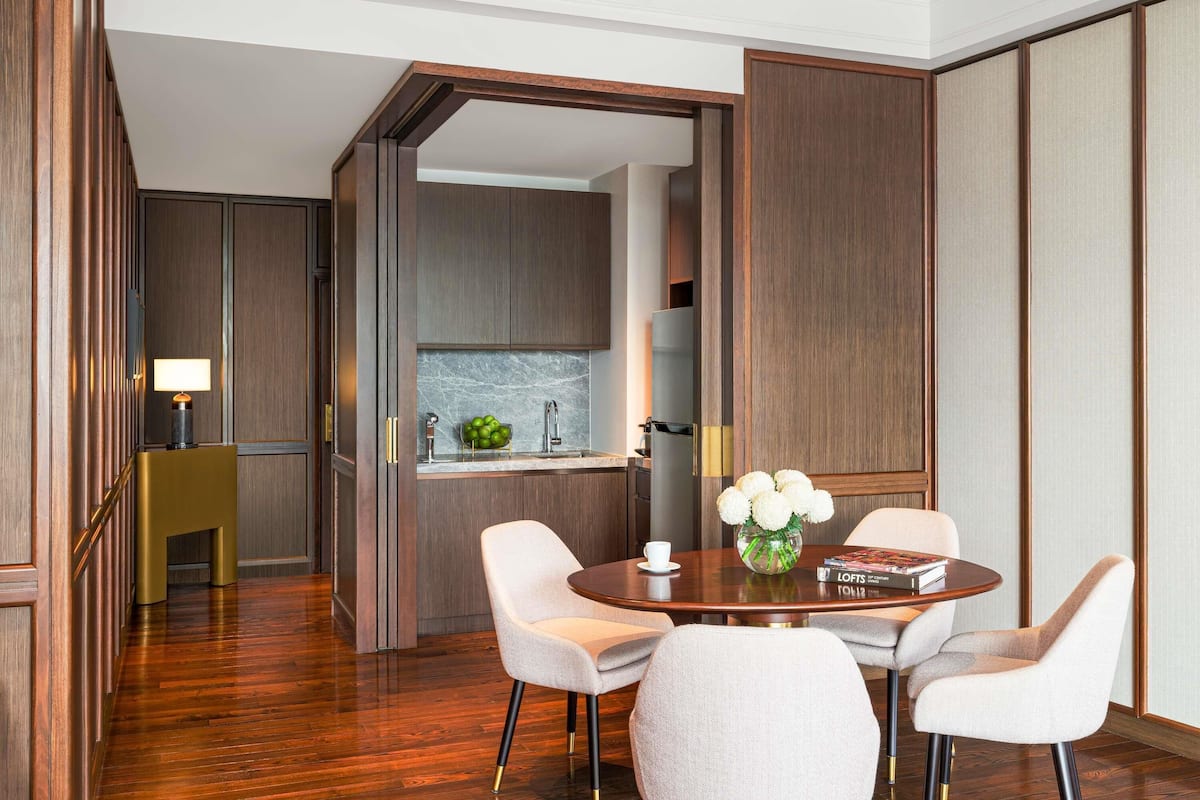 A modern hotel room with a wooden floor, a dining area with a round table and four chairs, a kitchen with a sink and cabinets, and a view through an open door.