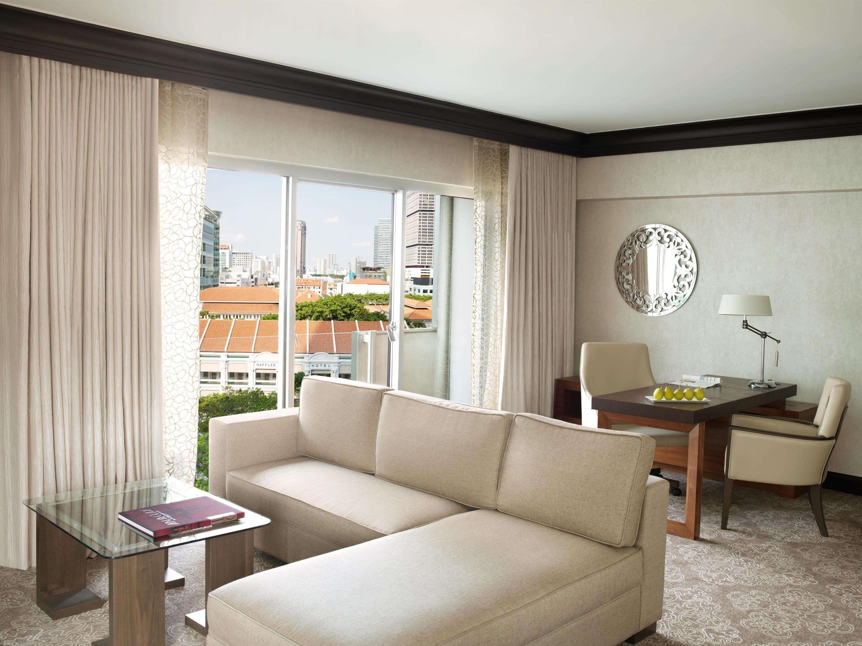 A hotel room with a sofa, a glass table with a book, a dining table with chairs, a lamp, and a view of the cityscape.