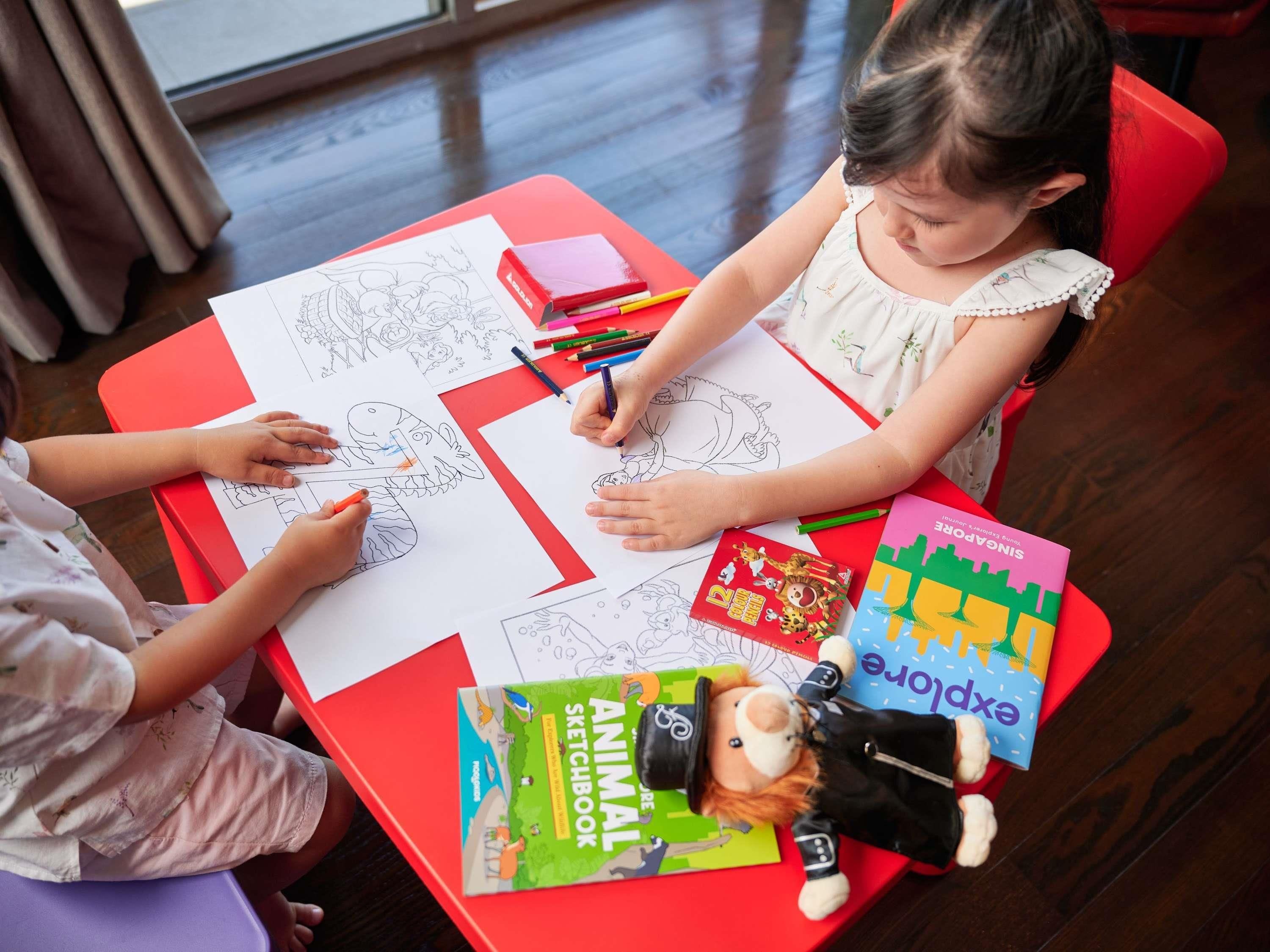 Two children drawing on large sheets of paper at a table with various coloring books and a stuffed toy.
