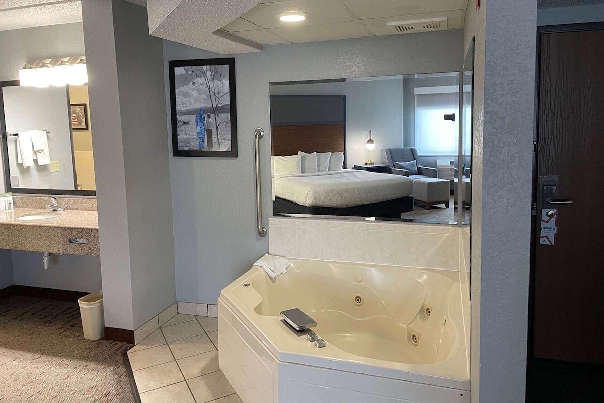 A hotel room with a large bathtub, a bed, a bedside table with a lamp, a mirror, and a bathroom with a sink and towel rack.