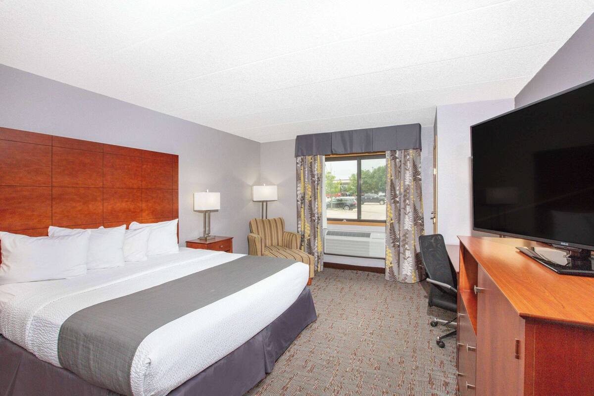 A hotel room with a large bed, a desk with a flat-screen TV, a chair, a lamp, and a window with curtains.