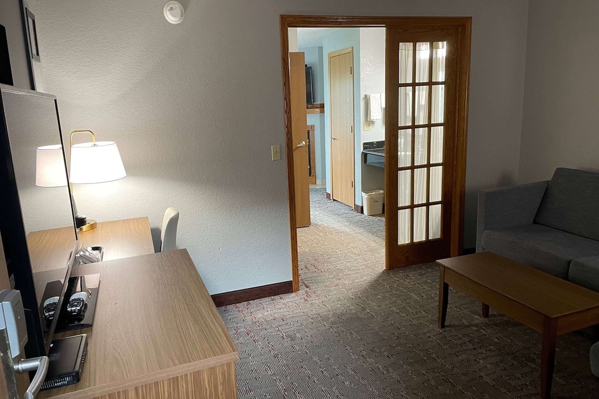 A hotel room with a wooden desk, a lamp, a sofa, and a view through a door to a bathroom and kitchen area.