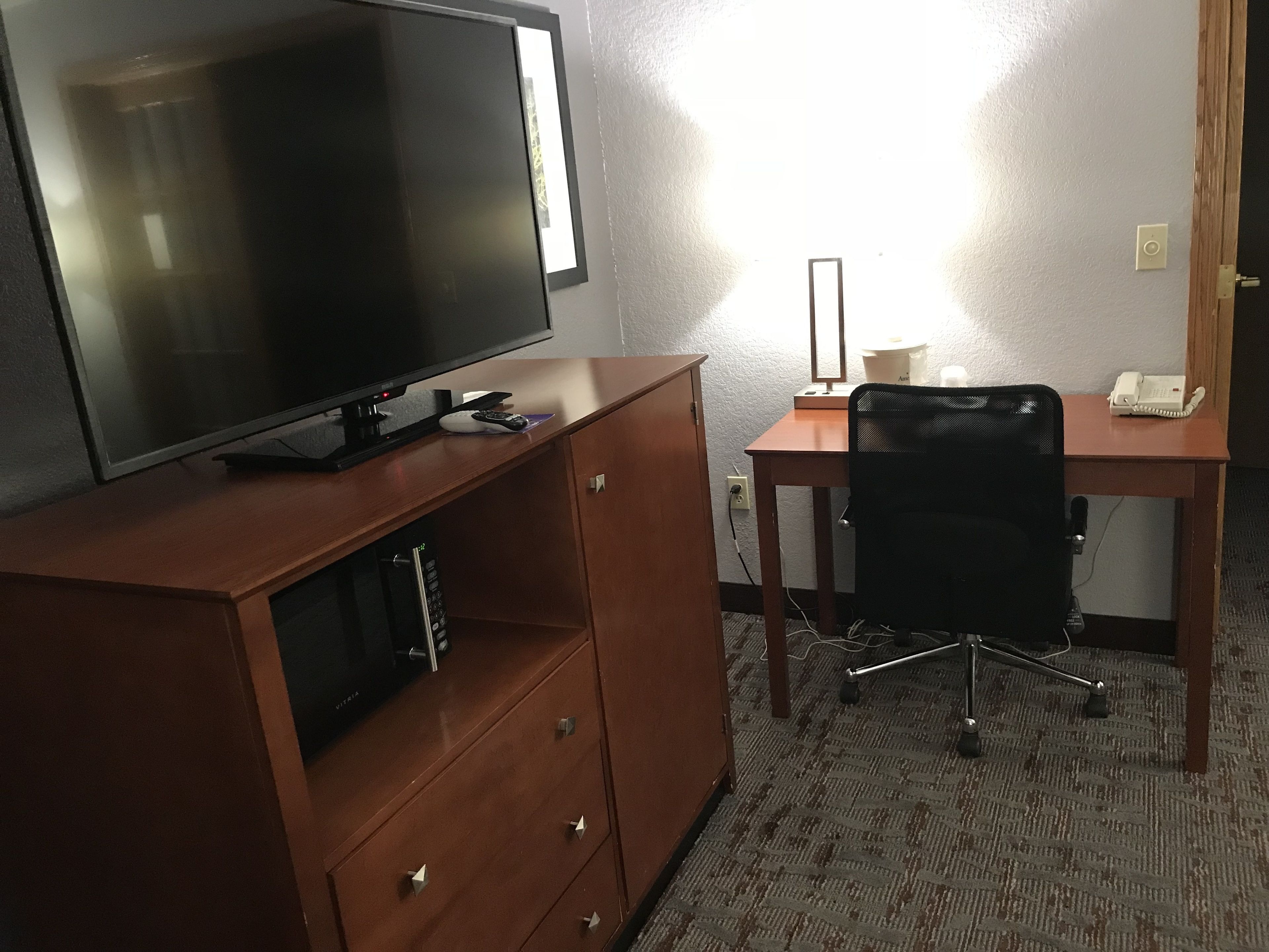 A hotel room with a flat-screen TV on a wooden cabinet, a desk with a chair, a telephone, and a lamp.