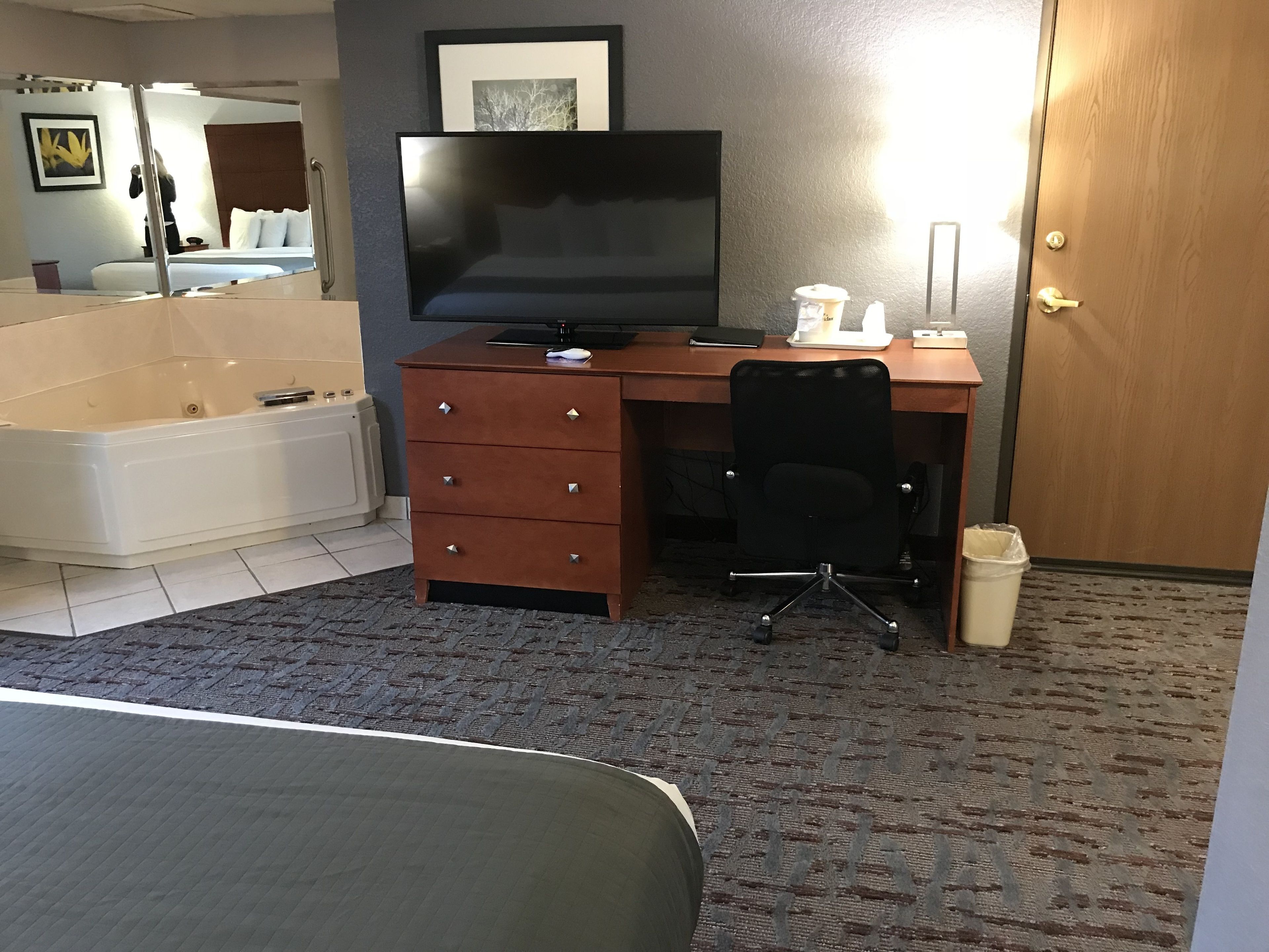 Ein Hotelzimmer mit einem großen Bett, einem Schreibtisch mit Computer, einem Sessel, einem Fernseher, einer Badewanne und einer Tür.