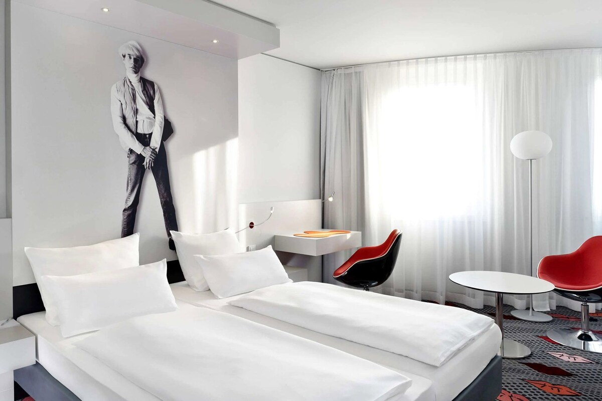 Een hotelkamer met een bed, een bureau, een stoel en een groot raam met gordijnen.