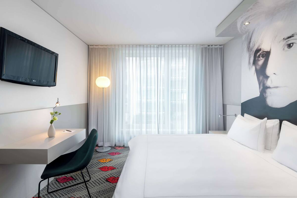 Une chambre d’hôtel comprenant un lit, un bureau avec une chaise, un téléviseur fixé au mur et une grande fenêtre avec des rideaux légers.