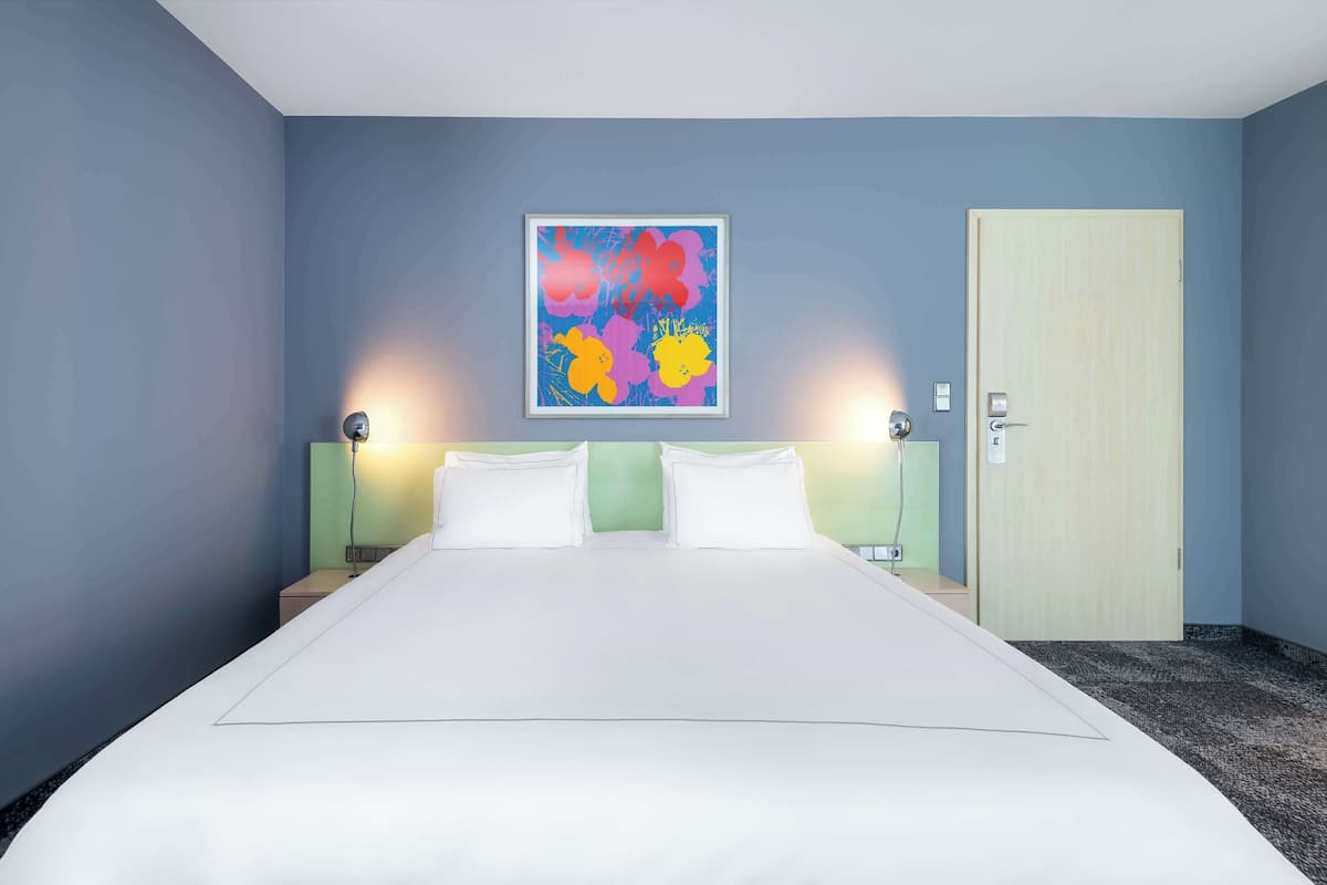 Une chambre d’hôtel avec un grand lit, deux tables de chevet, une commode, une œuvre d’art florale encadrée au mur et une porte.