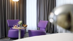 Hotelkamer met twee paarse fauteuils, een kleine tafel met een schaal fruit en een bloevas, plus een grote ronde lamp.