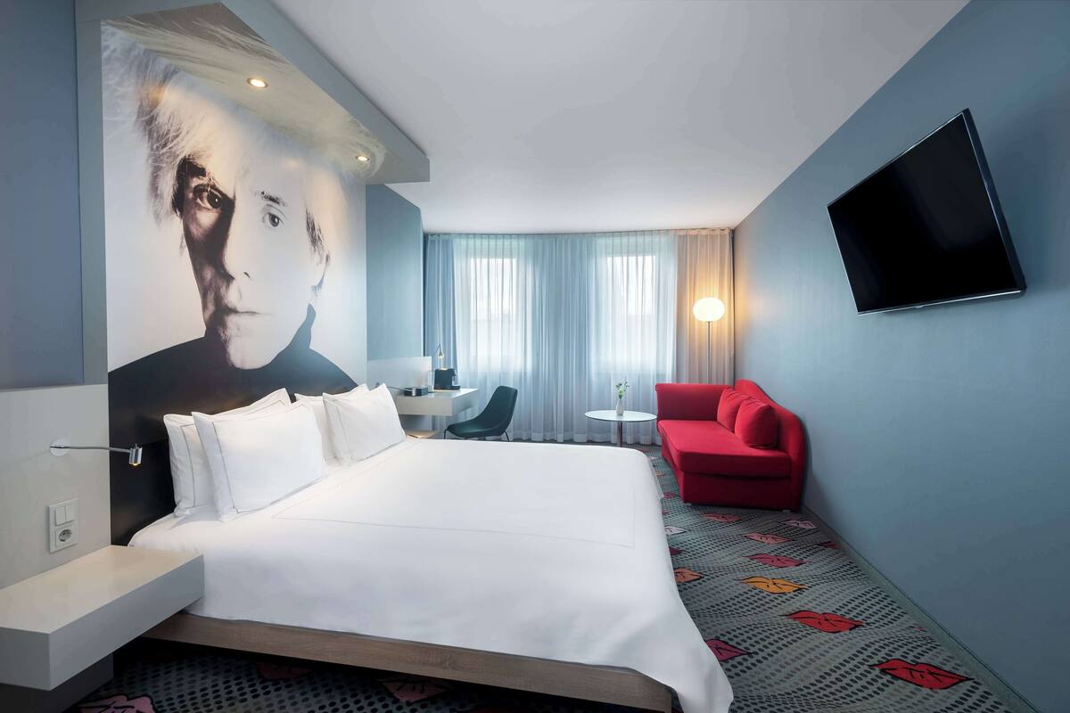 Une chambre d’hôtel avec un grand lit, un fauteuil rouge, une petite table et un téléviseur fixé au mur.