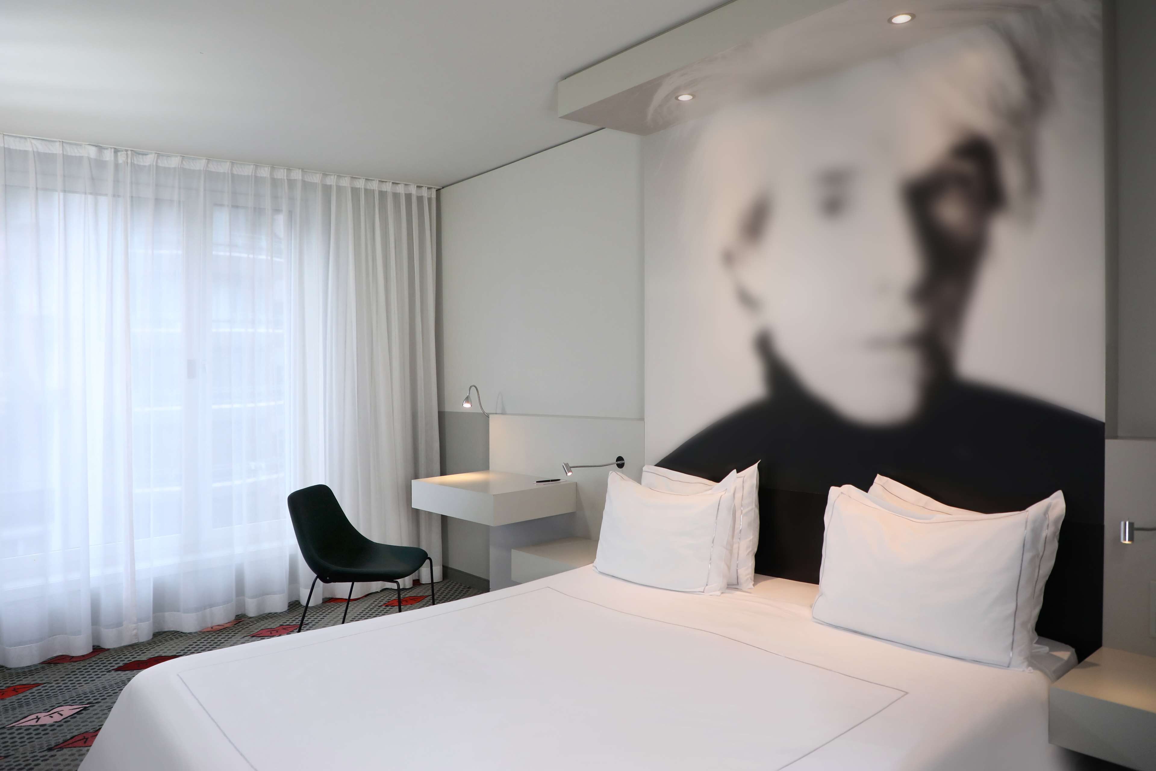 Una habitación de hotel moderna con una cama grande, una mesita de noche, una silla, un escritorio y una pantalla grande montada en la pared.